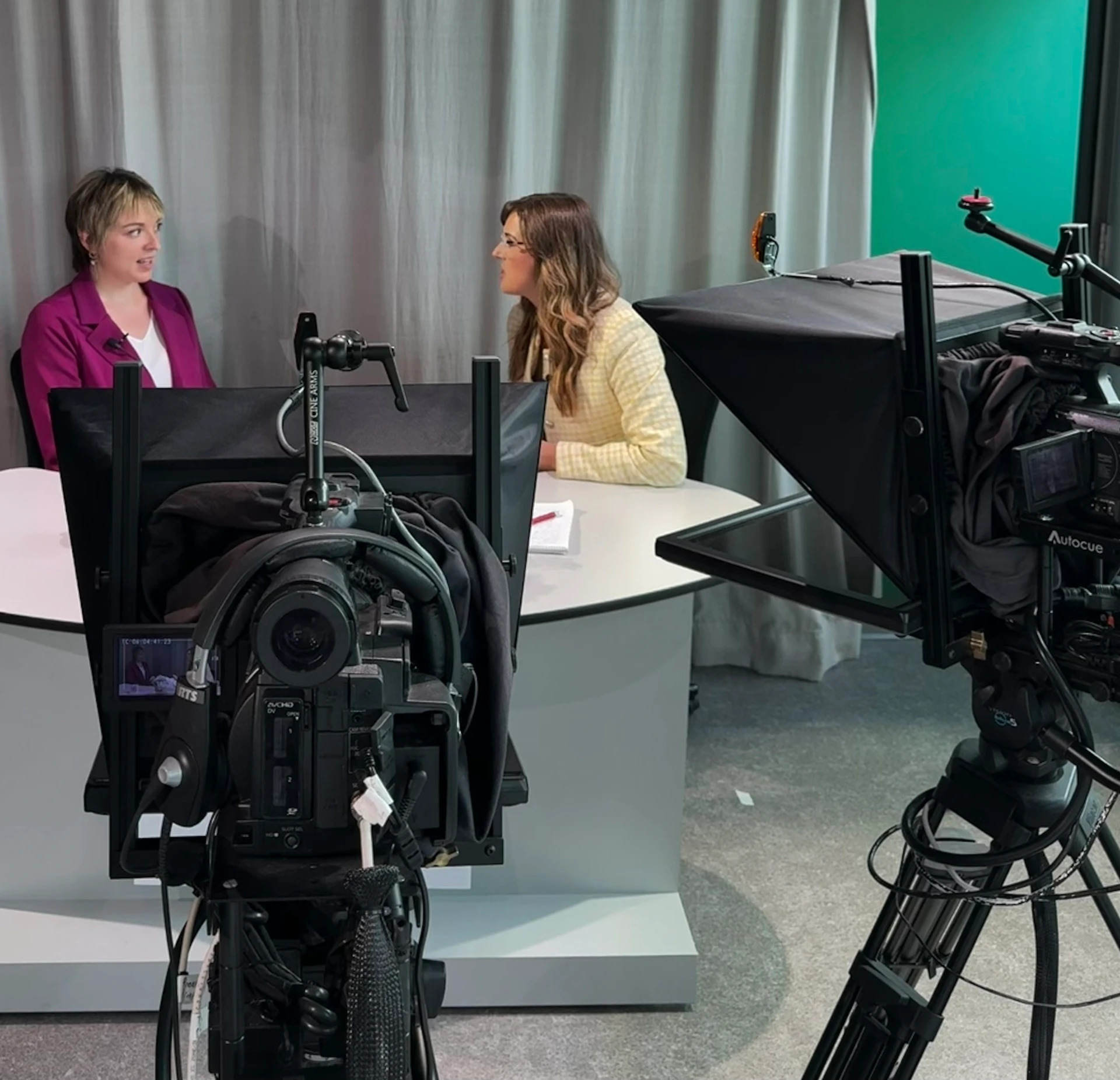 Two journalists in a modern broadcast studio interview, seen across cameras and a teleprompter